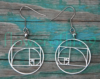 May include: A pair of silver earrings with a geometric design inspired by the Fibonacci sequence. The earrings are circular and feature a series of concentric circles and squares.