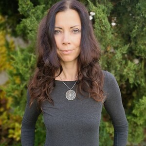 May include: A woman with long brown hair wearing a gray long-sleeved shirt and a silver necklace with a round pendant featuring a geometric design.