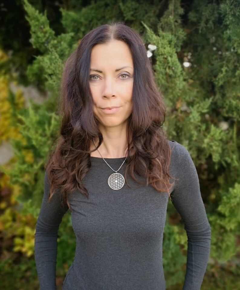 May include: A woman with long brown hair wearing a gray long-sleeved shirt and a silver necklace with a round pendant featuring a geometric design.