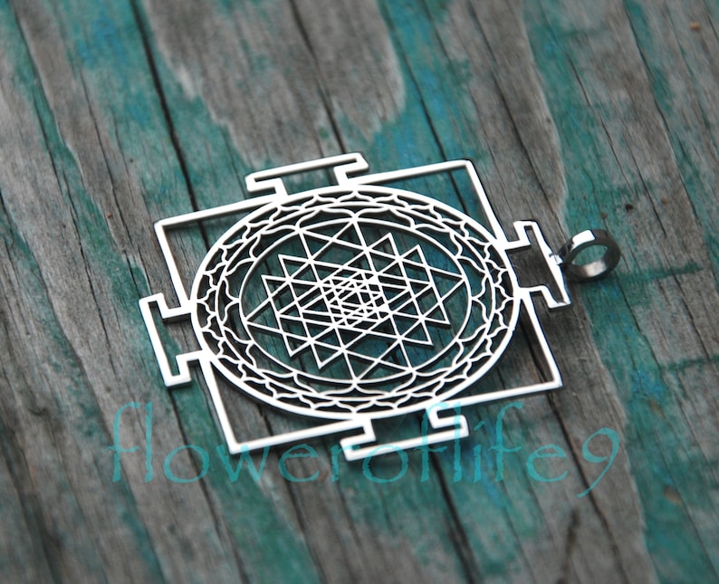 May include: A silver pendant featuring a Sri Yantra design, a sacred geometric symbol in Hinduism. The pendant is intricately detailed and has a circular shape with a square frame.