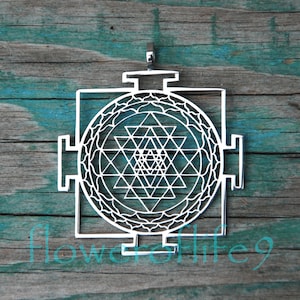 May include: A silver pendant featuring a Sri Yantra, a sacred geometric symbol in Hinduism, with intricate details and a circular design. The pendant is on a wooden background.