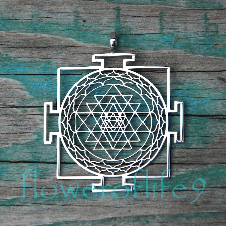 May include: A silver pendant featuring a Sri Yantra, a sacred geometric symbol in Hinduism, with intricate details and a circular design. The pendant is on a wooden background.