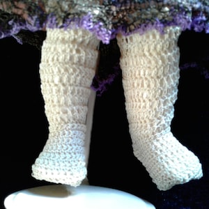 May include: A pair of white crocheted leg warmers with a lacy texture. The leg warmers are worn by a doll and are displayed against a black background.