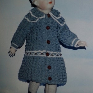 May include: A porcelain doll wearing a blue knitted dress with white trim and brown buttons. The doll is wearing a white knitted hat with a pink bow. The doll is standing on a light blue background.