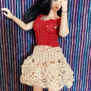 May include: A doll wearing a handmade red crochet top and a cream-colored crochet skirt. The doll has long black hair and is posed against a striped background with shades of blue, pink, and purple. The crochet work appears delicate and detailed.