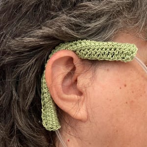 May include: A light green crocheted ear protector designed to cushion the ears from the pressure of oxygen tubing. The handmade accessory is designed to provide comfort and support for individuals using nasal cannulas. The soft, textured design is visible.