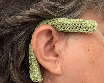May include: A sage green crochet ear cushion cover designed to fit over the tubing of a nasal cannula. The cover features a textured, hand-stitched pattern that wraps around the plastic tubing to provide a soft layer between the tube and skin.