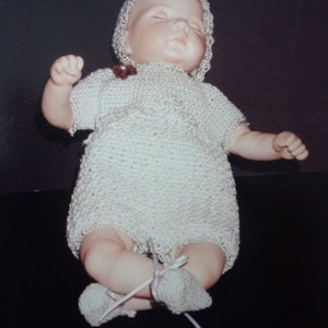 May include: A white crocheted baby doll wearing a white crocheted bonnet and booties. The doll is lying on its back with its eyes closed.