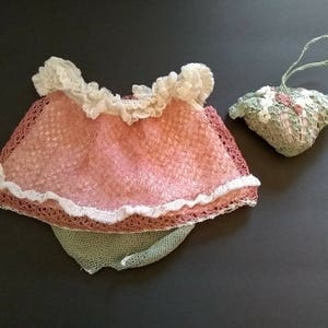 May include: A vintage-style, handmade set of baby clothes, featuring a pink and white lace top and a matching green bottom. A small, decorative, lace-covered pouch with a handle is also included. The items are displayed on a dark gray surface.