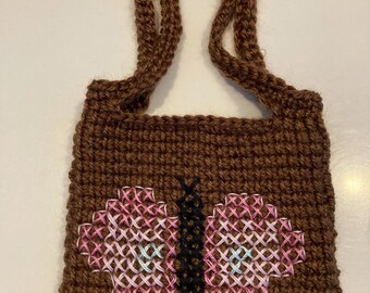 May include: A brown crocheted tote bag with a butterfly design. The butterfly has pink, white, and light blue wings and a black body. The bag has a handle made of the same brown yarn.