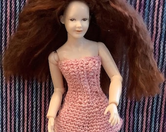 May include: A doll with long, wavy auburn hair and a pale complexion, wearing a sleeveless, light pink, hand-knitted dress. The doll features articulated joints at the shoulders and elbows, with a neutral facial expression and light brown eyes.