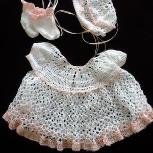 May include: A white crocheted baby dress with a pink trim, a matching white crocheted hat with a pink trim, and a pair of white crocheted booties with a pink trim.