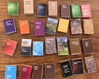 Op de afbeelding: Een verzameling miniatuurboeken met titels over huisverbetering, houtbewerking en design. De boeken zijn in verschillende kleuren en maten, gerangschikt op een houten oppervlak. Titels zijn onder meer "Hoe je een huis bouwt" en "Hout begrijpen".