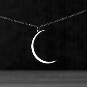 Op de afbeelding: Een zilveren maanvormige hanger ketting. De hanger heeft een eenvoudige, gebogen vorm en hangt aan een fijne ketting. De achtergrond is donker en effen, wat de heldere, metalen glans van de ketting benadrukt.