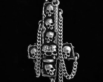 May include: A silver pendant necklace with a large, ornate cross pendant featuring multiple skull designs. The cross is adorned with chains and has a dangling chain at the bottom.