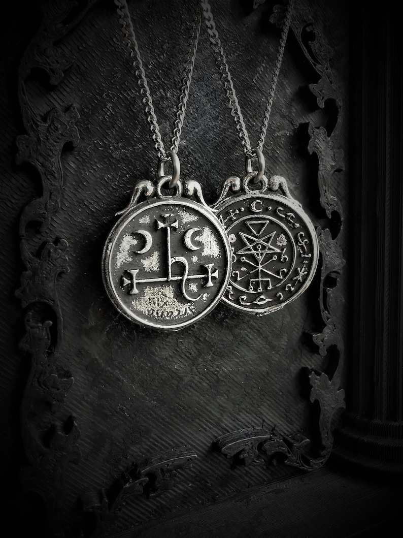 May include: Two silver necklaces with pendants featuring sigils. The pendant on the left has a crescent moon, a cross, and a symbol that resembles a letter L. The pendant on the right has a pentagram, a triangle, and other symbols. The necklaces are hanging against a black background.