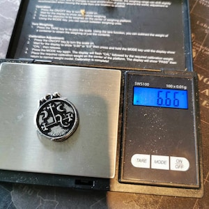 May include: A silver pendant with a black design of a pentagram and other symbols is on a digital scale. The scale reads 6.66 grams. The scale is labeled "SWS100" and has a tare, mode, and on/off button.