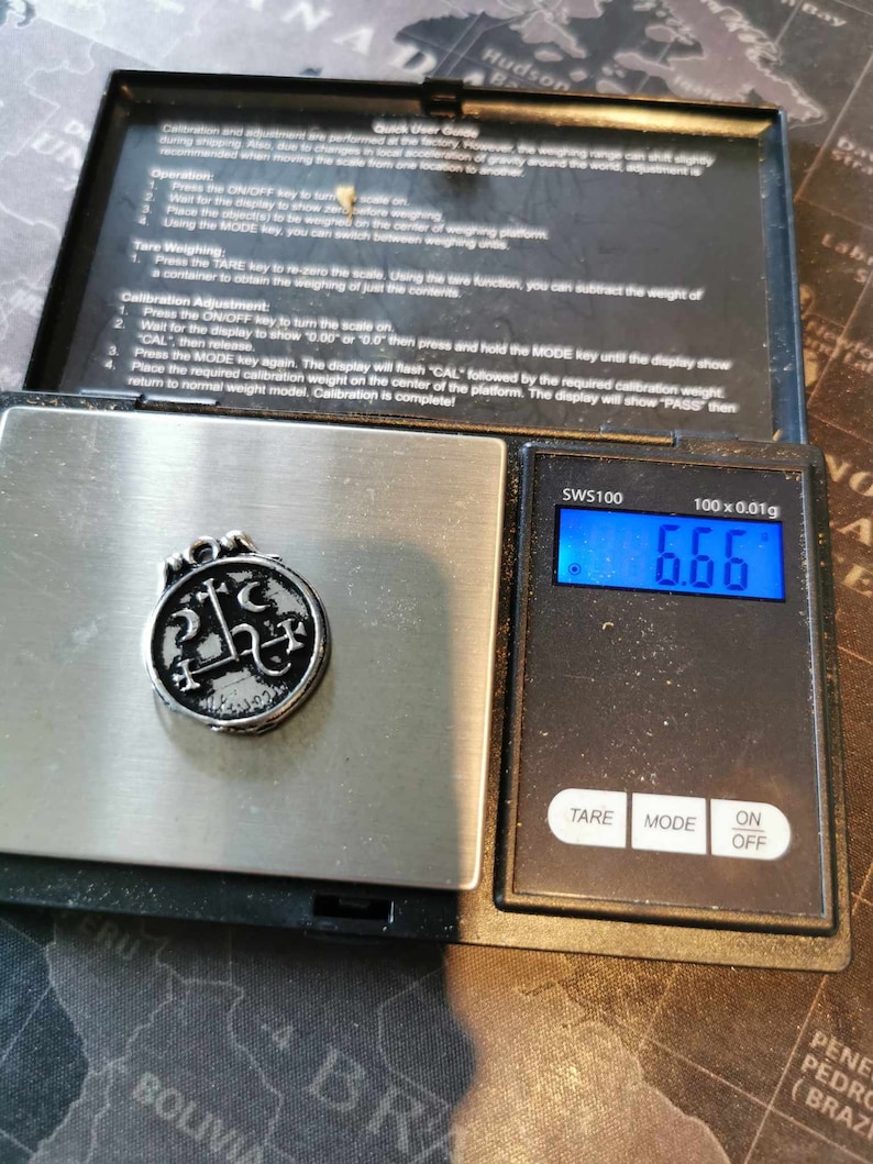 May include: A silver pendant with a black design of a pentagram and other symbols is on a digital scale. The scale reads 6.66 grams. The scale is labeled "SWS100" and has a tare, mode, and on/off button.