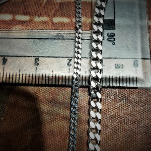 May include: Two silver chains, one with a more traditional curb link design and the other with a flat, wider link design, are laid out on a brown surface next to a ruler.