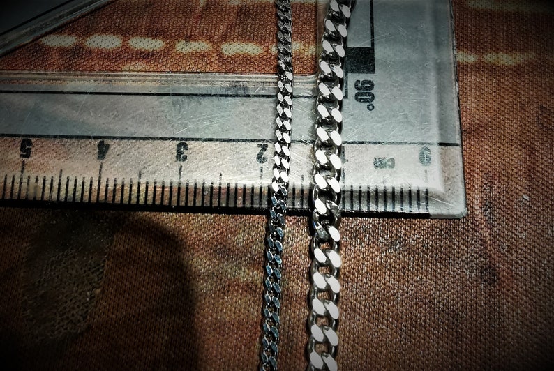 May include: Two silver chains, one with a more traditional curb link design and the other with a flat, wider link design, are laid out on a brown surface next to a ruler.