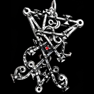 May include: A silver pendant with a complex, geometric pattern. It has a central red stone and a loop for suspension. The background is a plain black, accentuating the pendant's design.