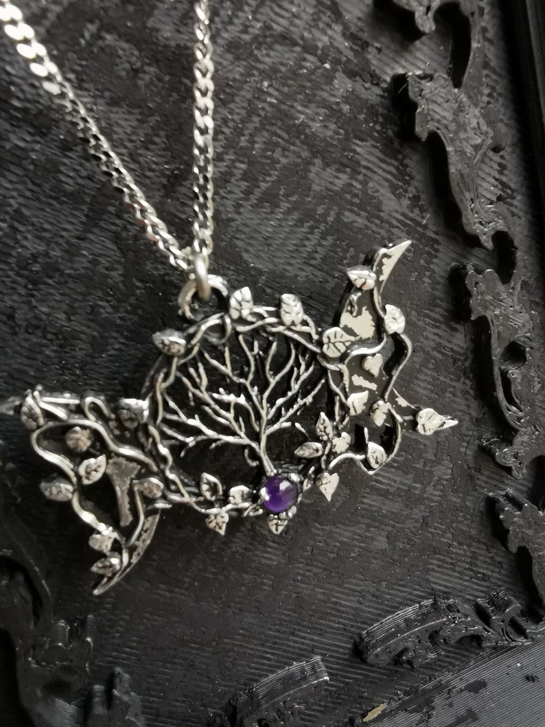 May include: A silver necklace with a pendant featuring a detailed tree of life design with leaves and a purple gemstone in the center.