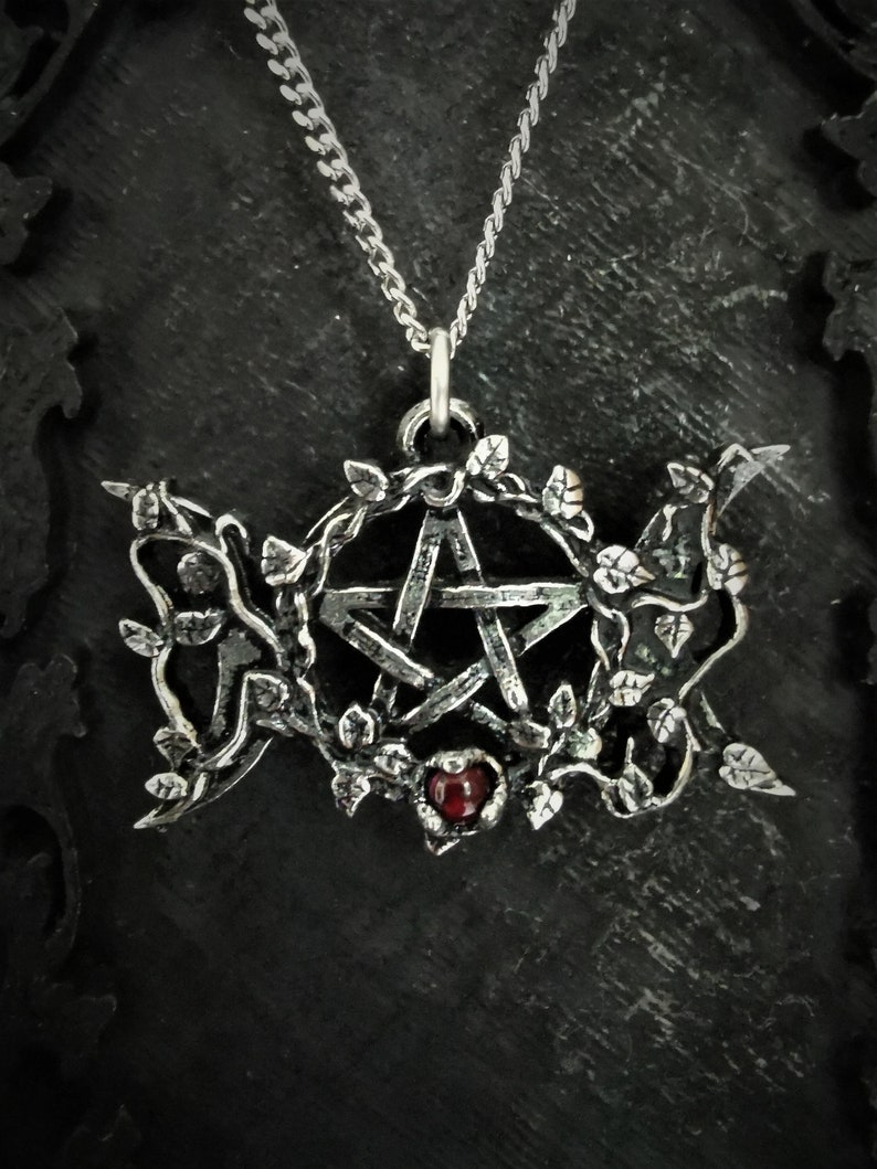 May include: A silver pendant necklace featuring a pentagram surrounded by vines and a red gemstone. The necklace is on a silver chain.