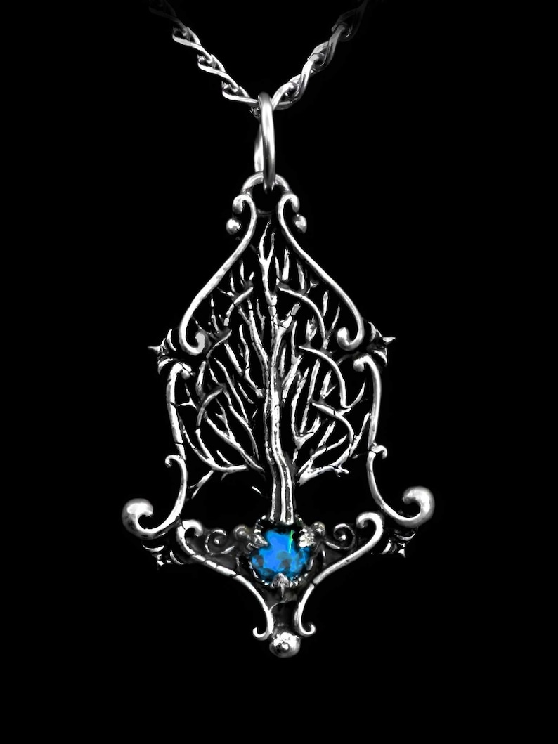 K&ouml;nnte beinhalten: Eine silberne Halskette mit einem detaillierten Baumdesign und einem blauen Edelstein an der Basis. Der Anh&auml;nger ist an einer silbernen Kette befestigt.