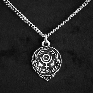 Könnte beinhalten: Eine silberne Halskette mit einem runden Anhänger, der ein stilisiertes Symbol eines Planeten mit einem Ring um ihn herum zeigt, umgeben von einem Kranz aus Blättern.