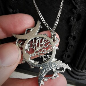 May include: Silver necklace with a tree of life pendant. The pendant is surrounded by two crescent moons and has the word "Arcana" inscribed on it.