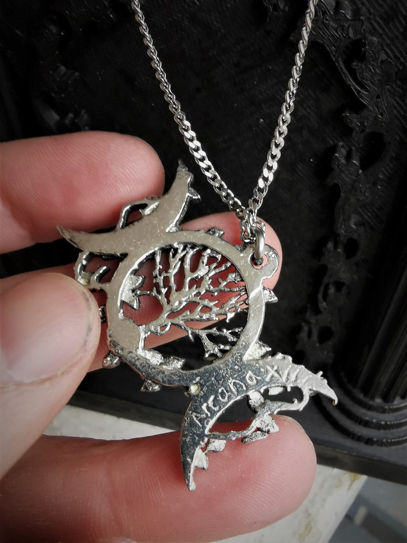 May include: Silver necklace with a tree of life pendant. The pendant is surrounded by two crescent moons and has the word "Arcana" inscribed on it.