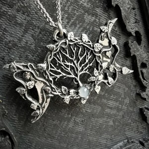 May include: A silver pendant necklace featuring a tree of life design with a moon and leaves. The pendant has a clear gemstone in the center.