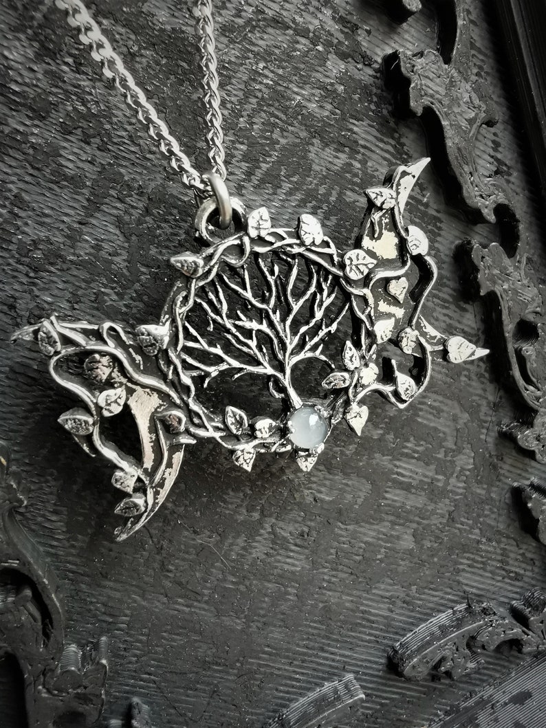 May include: A silver pendant necklace featuring a tree of life design with a moon and leaves. The pendant has a clear gemstone in the center.