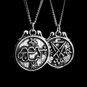 Op de afbeelding: Twee zilverkleurige kettingen met hangers. Elke ronde hanger toont gedetailleerde symbolen, waaronder sterren en geometrische vormen. De kettingen hangen tegen een zwarte achtergrond, wat de metalen glans benadrukt.