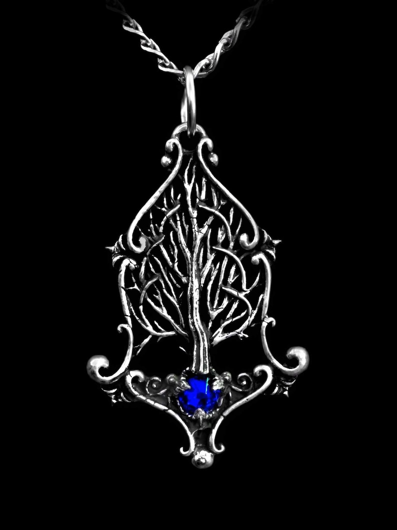 K&ouml;nnte beinhalten: Eine silberne Halskette mit einem Anh&auml;nger, der ein detailliertes Baumdesign mit einem blauen Edelstein an der Basis zeigt. Der Baum ist von filigranen Schn&ouml;rkeln umgeben.