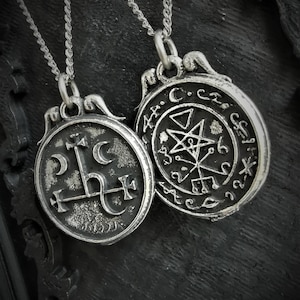 May include: Two silver necklaces with round pendants. The pendant on the left features a stylized symbol with a cross and crescent moon. The pendant on the right features a pentagram and other symbols.