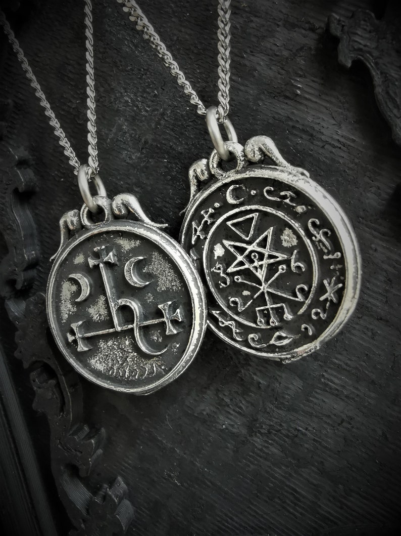 May include: Two silver necklaces with round pendants. The pendant on the left features a stylized symbol with a cross and crescent moon. The pendant on the right features a pentagram and other symbols.