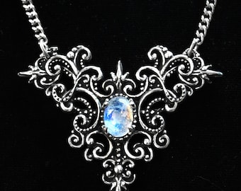 May include: A silver necklace with a large, ornate pendant featuring a moonstone. The pendant is made of intricate filigree work and has a delicate chain.