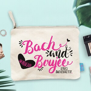 May include: A white canvas makeup bag with a black zipper. The bag has a pink and black design that says "Bach and Boyee" with a heart that says "San Diego 2022". The text below the design says "Leila's Bachelorette."