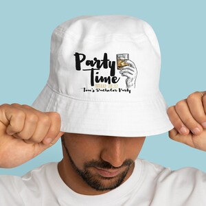 Op de afbeelding: Witte vissershoed met de tekst "Party Time" in zwart schrift, een illustratie van een hand die een glas whisky vasthoudt en de tekst "Tom's Bachelor Party". Een casual accessoire.