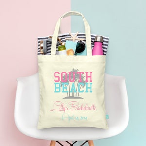 May include: A white canvas tote bag with the text "South Beach" in pink and blue lettering. The bag also has the text "Lilly's Bachelorette April 18, 2014" in gray lettering. The bag is filled with various items, including a beach towel, sunglasses, a water bottle, and a seashell.