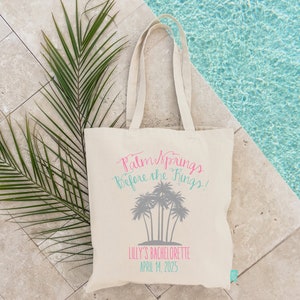 May include: A white canvas tote bag with the text "Palm Springs Before the Rings!" in pink and teal lettering. The bag also features a graphic of palm trees and the text "Lilly's Bachelorette April 14, 2025" in pink lettering.
