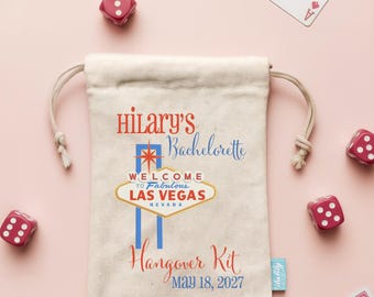 Könnte beinhalten: Ein cremefarbener Beutel mit Kordelzug, der den Text "Hilary's Bachelorette Hangover Kit" und "MAY 18, 2027" sowie eine Grafik des "Welcome to Fabulous Las Vegas"-Schildes zeigt, umgeben von verstreuten Spielkarten und roten Würfeln.