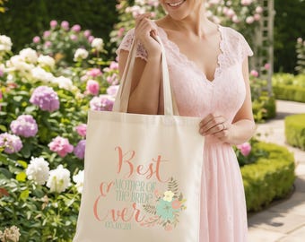 May include: A cream-coloured canvas tote bag featuring the text "Best MOTHER OF THE BRIDE Ever" in pink and teal script and block lettering, accompanied by a floral graphic and the date "05.18.28" printed in a simple font at the bottom.