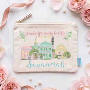 May include: A beige canvas zip-up pouch featuring a colourful illustration of the Savannah city skyline. The text reads Lindsey's Bachelorette July 2028 and Savannah. The design includes pink, green, and yellow buildings with a gold zip pull.