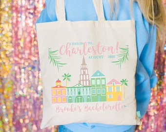 Könnte beinhalten: Eine weiße Canvas-Tragetasche mit einer rosa, grünen und gelben Illustration von Charleston, South Carolina. Der Text "Cheers to... Charleston! August 2026" ist in rosa und grün gedruckt. Der Text "Brooke's Bachelorette" ist in rosa am unteren Rand der Tasche gedruckt.