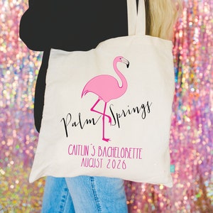 May include: A white canvas tote bag with a pink flamingo illustration and the text "Palm Springs" in black script. The bag also has the text "Caitlin's Bachelorette August 2026" printed on it.