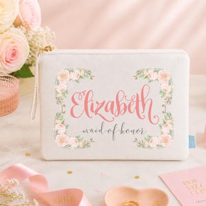 May include: A rectangular white zippered pouch featuring a floral border of pink roses and green leaves. The text "Elizabeth" is printed in large pink script, with "maid of honour" written below in smaller black cursive lettering.