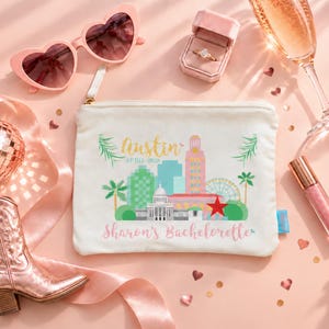 May include: A white canvas zip-up pouch featuring a colourful Austin city skyline graphic with the text Austin April 2025 and Sharon's Bachelorette. The pouch is surrounded by heart-shaped sunglasses, a ring in a box, a champagne flute, and a boot.