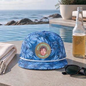 Op de afbeelding: Een blauwe honkbalpet met een palmbladpatroon, een wit koord en een ronde beige patch aan de voorzijde. De patch bevat een portret van een persoon en de tekst "JONH'S BACHELOR PARTY DRINKING TEAM" in een cirkelvormige opstelling.